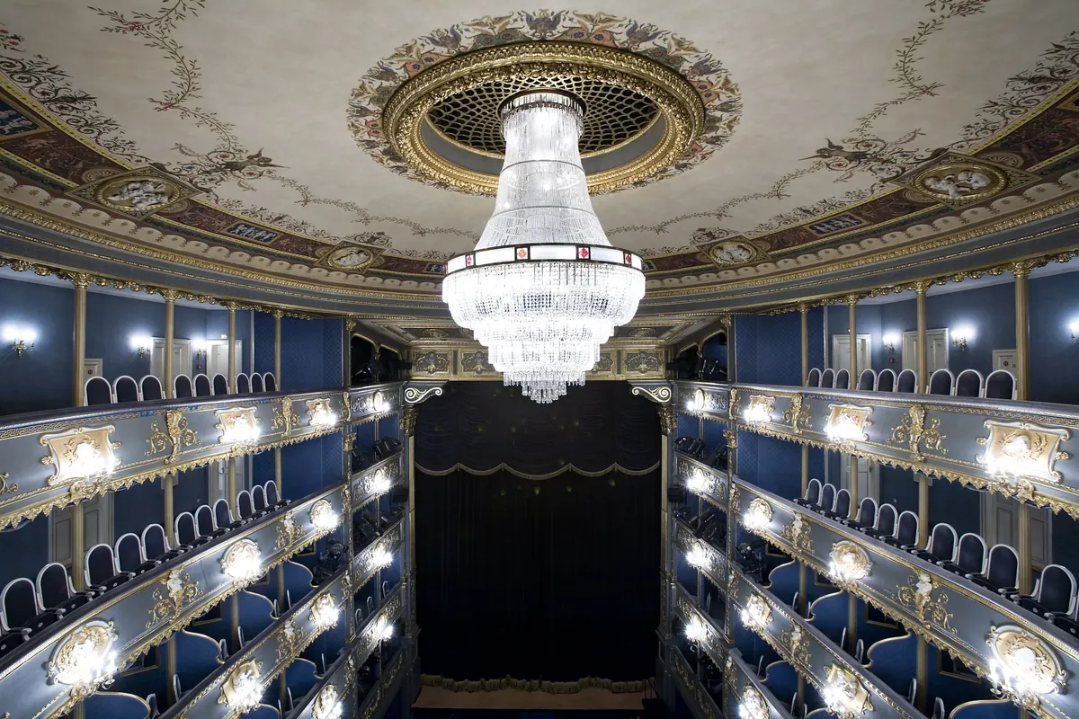 The tiered gilded boxes and crystal chandelier of the Estates Theatre in Prague, seen from the stalls toward the stage curtain.
