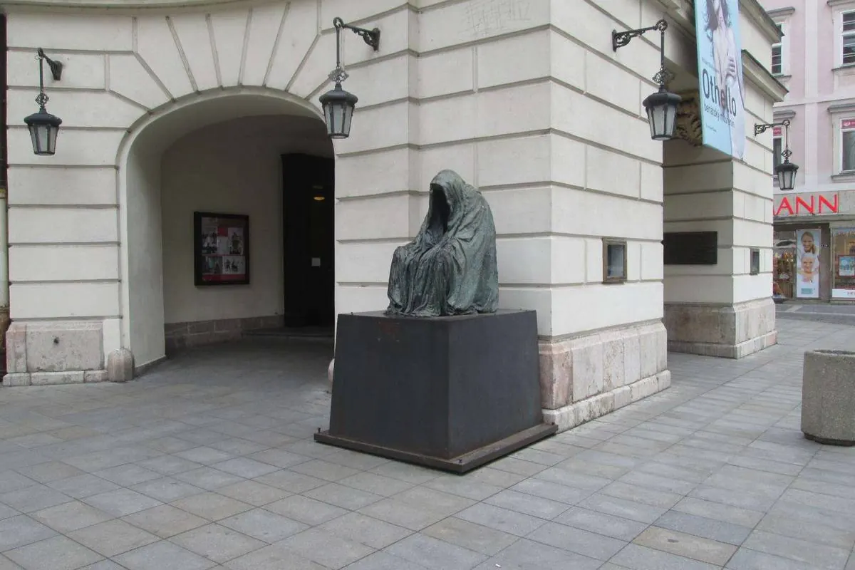 A bronze statue of a hooded, faceless figure on a dark plinth under an arched white wall, outside the Estates Theatre.