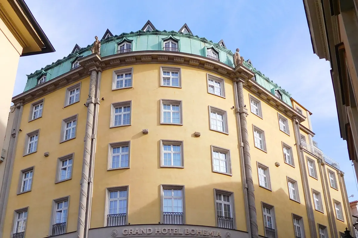 The curved yellow corner facade of the Grand Hotel Bohemia, a six-storey building with a green roof, on a Prague street.